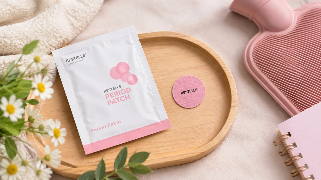 Pink Restelle period patch and sachet resting on a wooden tray with a hot water bottle, chamomile flowers, and notebook